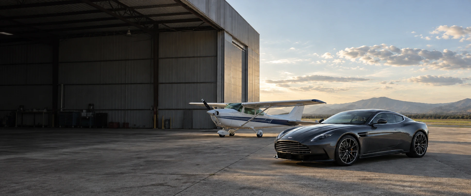 A polished graphite sports car and small white high-wing propeller plane together outside a hangar
